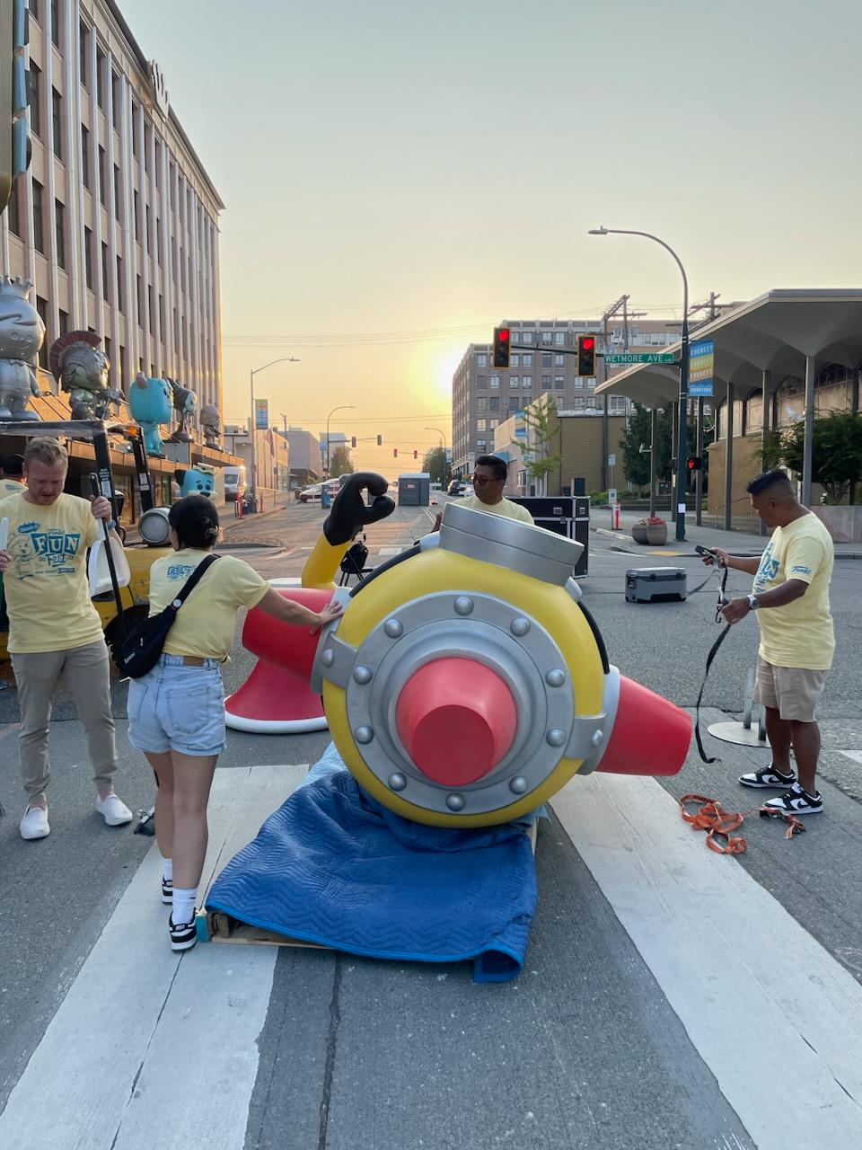 Minion Statue: Funko volunteers in yellow Fun Fest 2023 tee shirts help place a Minion statue out in the street.